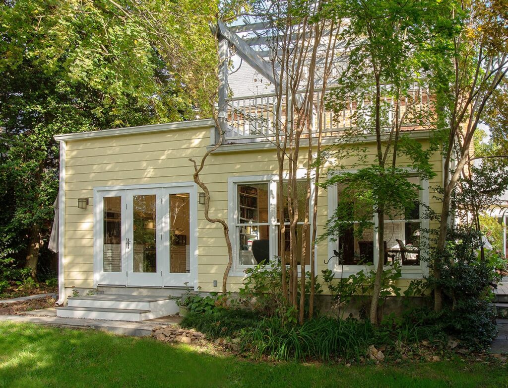 Rear view of a two-story home in the American University Park neighborhood of Washington, DC, featuring a newly built first-floor addition by Spectrum Design Build. The addition extends from the back of the house with a modern design that blends with the existing structure. The exterior features neutral-colored yellow siding wit bright outdoor greenery,